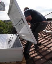 Hombre instalando paneles solares en un tejado inclinado de tejas marrones.