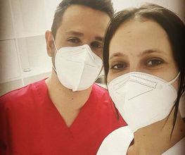 Dos personas con mascarillas, una con uniforme rojo y otra con blanco, en un entorno clínico.