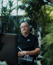 Ein Mann sitzt in einem sonnigen Raum voller Pflanzen vor einem Computer mit Kamerabeleuchtung.