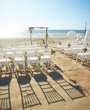 Ceremonia de boda en la playa con sillas blancas, alfombra y letras grandes bajo el sol.