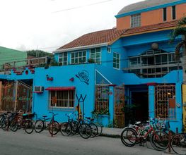 Edificio azul con bicicletas estacionadas al frente, rodeado de vegetación y cielos nublados.