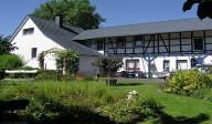 Weißes Fachwerkhaus mit Garten, von Bäumen umgeben, sonniger Tag.