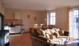 Wohnzimmer mit braunem Sofa, Esstisch, Küche, heller Wand, großen Fenstern und Vorhängen.