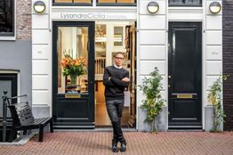 Een persoon poseert voor de ingang van een stijlvolle kapperszaak met bloemdecoratie.