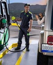 Empleado de gasolinera atendiendo a cliente en un Jeep dorado. Montañas al fondo.