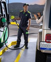 Trabajador de gasolinera llenando un vehículo mientras una mujer sonríe desde el auto.