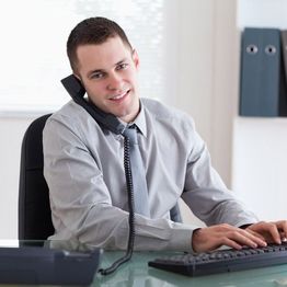 Man at a desk on the phone, smiling, typing on a keyboard, office setting.