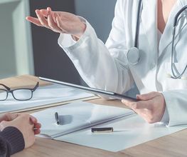 Ein Arzt mit Stethoskop spricht mit einem Patienten. Auf dem Tisch liegen Unterlagen und eine Brille.