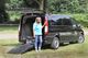 Frau mit blauem Shirt steht an einem schwarzen Fahrdienst-Van mit geöffneter Heckklappe und Rampe.