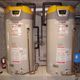 Two propane water heaters with digital displays, pipes, and safety labels in a utility room.
