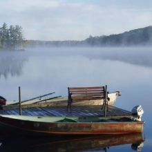 A foggy lake scene with two small boats moored to a wooden dock and trees in the background.