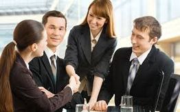 Three people sitting around a table while a woman shakes hands with one of them.
