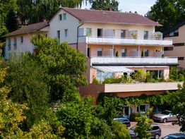 Mehrstöckiges Haus mit Balkonen, umgeben von üppigen Bäumen und parkenden Autos davor.