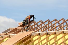 Worker installing wooden roof trusses on a building under clear blue sky.