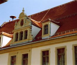 Altes Gebäude mit rotem Satteldach, weißen Fassaden und dekorativen Fenstern unter blauem Himmel.