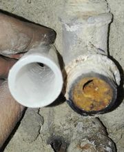 Mano sosteniendo una tubería plástica junto a una tubería de metal oxidada en una pared.