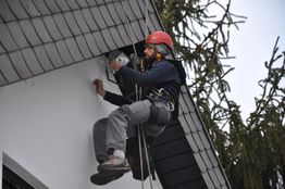 Ein Mann klettert gesichert an der Hauswand, trägt Helm und arbeitet an einem Gerät.