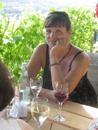 Frau sitzt an einem Tisch im Freien mit Weingläsern und lächelt in Richtung Kamera.