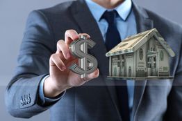 Businessman holding a dollar sign near a house covered in money, symbolizing real estate finance.