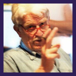 Älterer Mann mit Brille und Schnurrbart zeigt mit zwei Fingern, trägt ein Jeanshemd.