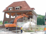 Bagger reißt ein zweistöckiges Haus mit rotem Dach ab, Arbeiter beobachten die Szene.