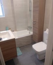 Baño moderno con lavabo, inodoro, bañera y mueble de madera. Luz natural por una ventana.