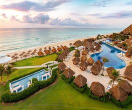 Vista aérea de un resort junto a la playa con piscinas y palapas al amanecer.