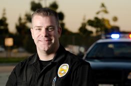 Police officer smiling outdoors with patrol car in the background at dusk.