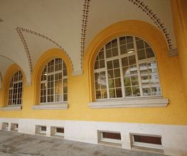Gelber Gebäudebogen mit großen Fenstern, weißer Simse und Verzierungen an der Decke.