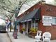 Brick building with canopy reads "Carpet & Blinds"; chairs and signs outside; tree in bloom nearby.
