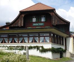 Altes Gasthaus mit Holzfassade und roten Dachziegeln, umgeben von grüner Landschaft, Himmel bewölkt.