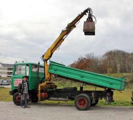Ein Mann steht neben einem grünen Kranlastwagen mit angehobenem Kipper auf einer Wiese.