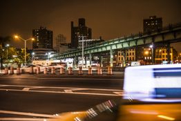 Bybillede om natten med bevægelige biler, en bro, bygninger og orange trafikstøtter.