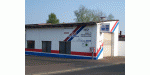 Lagerhalle mit diagonalen blauen und roten Streifen auf weißer Wand, umgeben von Bäumen.