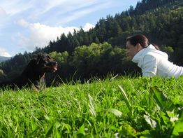 Frau in weißem Pullover liegt auf Wiese, schaut Hund an, Wald und Himmel im Hintergrund.