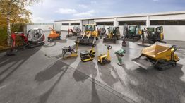 Baumaschinen vor einem Gebäude aufgestellt, darunter Bagger, Walze und Rüttelplatte.