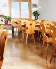 Gemütlicher Gastraum mit Holzstühlen und -tischen, Herzverzierungen und Pflanzen am Fenster.