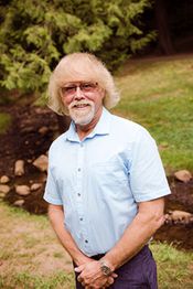 Man with white hair and beard wearing glasses, stands in a park with trees and rocks in the background.