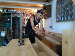 Mann in Werkstatt hobelt Holz auf Werkbank, im Hintergrund Regale und Werkzeuge.