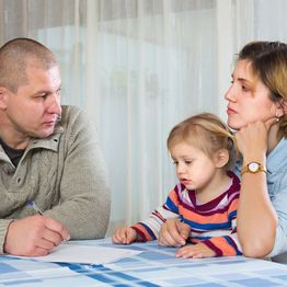 A family of three sits at a table with serious expressions, discussing paperwork.