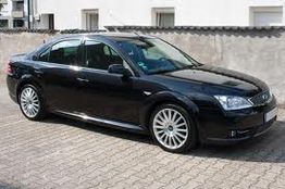 Schwarzes Auto, seitlich auf Pflastersteinen geparkt, vor einer weißen Wand.