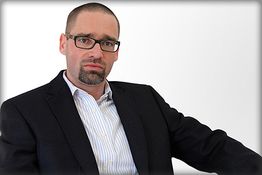 Eine ernst schauende Person mit Brille in einem dunklen Anzug vor weißem Hintergrund.