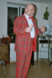 Mann im rot-karierten Anzug spricht lächelnd in Mikrofon in einem Raum mit Pflanzen.