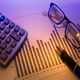 Calculator, glasses, and pen on paper with a bar graph under a purple light.
