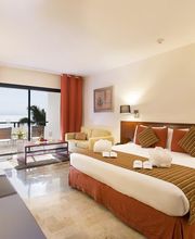 Habitación de hotel con cama grande, ventanal, sofá y tonos cálidos. Decoración moderna.