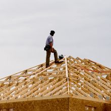 Construction worker atop wooden roof frame under a gray sky.