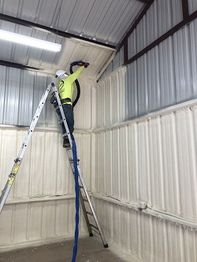 Worker on ladder applying spray foam insulation to a metal building interior wall.