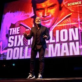 Man op podium, voor een scherm met "The Six Million Dollar Man" en felle kleuren.