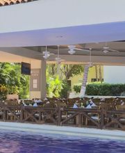 Restaurante al aire libre junto a una piscina, rodeado de vegetación y mesas dispuestas.