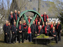 Gruppe von Menschen in Schwarz mit roten Akzenten posiert vor einem großen, grünen Rad im Freien.
